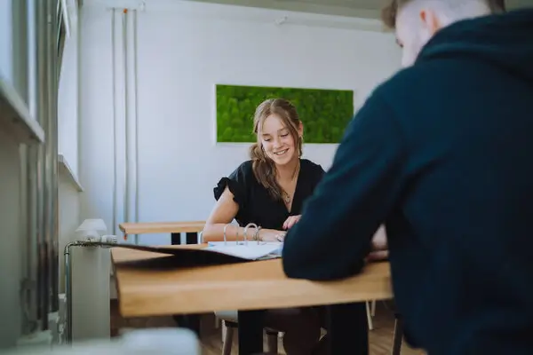 Lächelnde Kollegin sitzt am Tisch im Gespräch mit einem Kollegen.