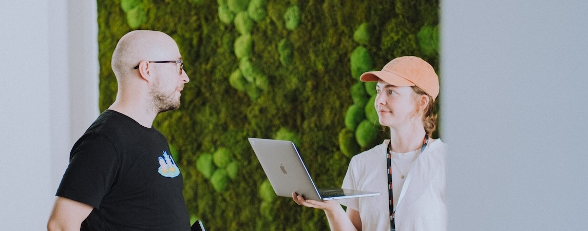 Zwei Kollegen mit Laptop vor grüner Wand.